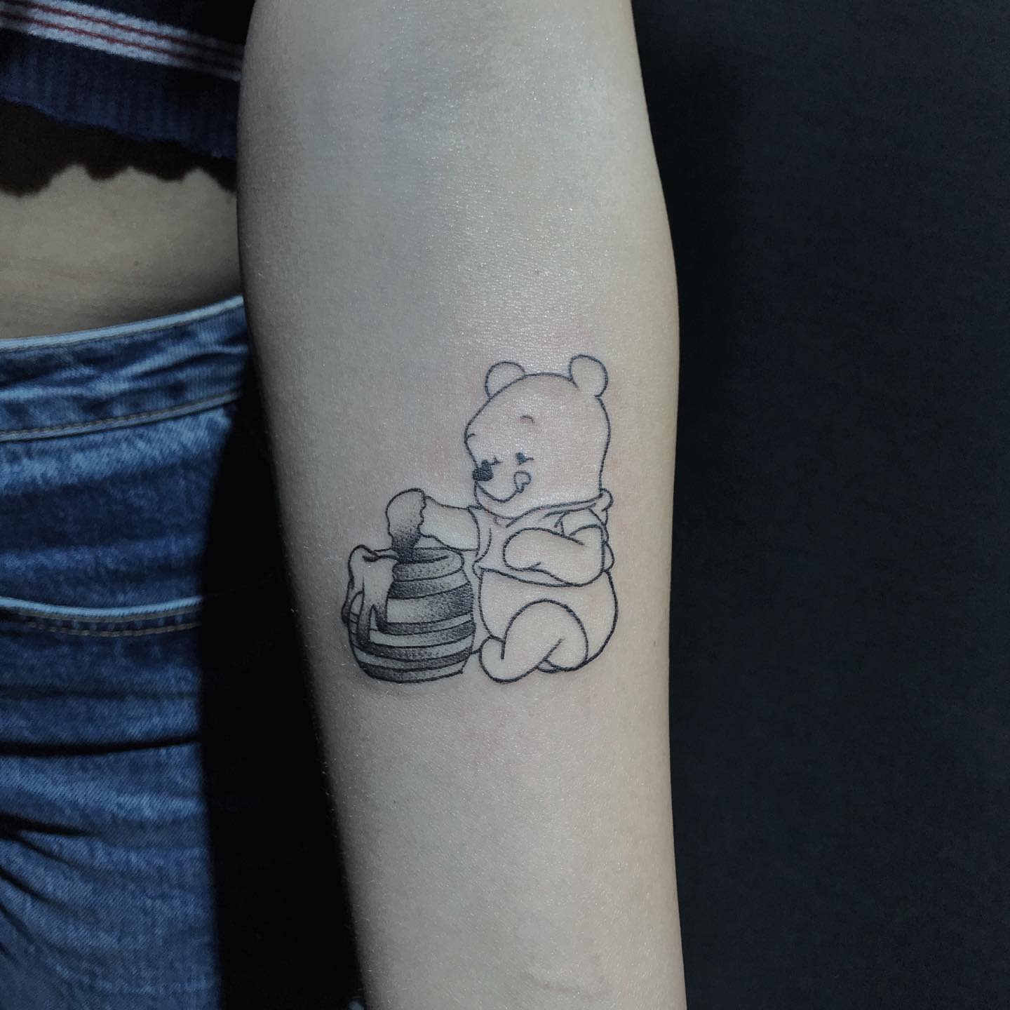 Simple black and white tattoo of Winnie the Pooh dipping his hand into a honey pot on a person's forearm